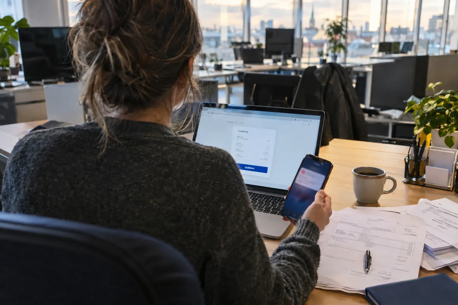 Illustration: en kontorsanställd sedd bakifrån vid ett skrivbord, på väg att godkänna en banköverföring, med en mobiltelefon som visar ett suddigt meddelande.
